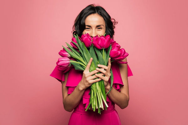 Eine fröhliche Frau in einem pinken Kleid hält einen Strauß pinker Tulpen vor ihrem Gesicht. Das Bild symbolisiert Stärke, Freude und Wertschätzung zum Weltfrauentag.